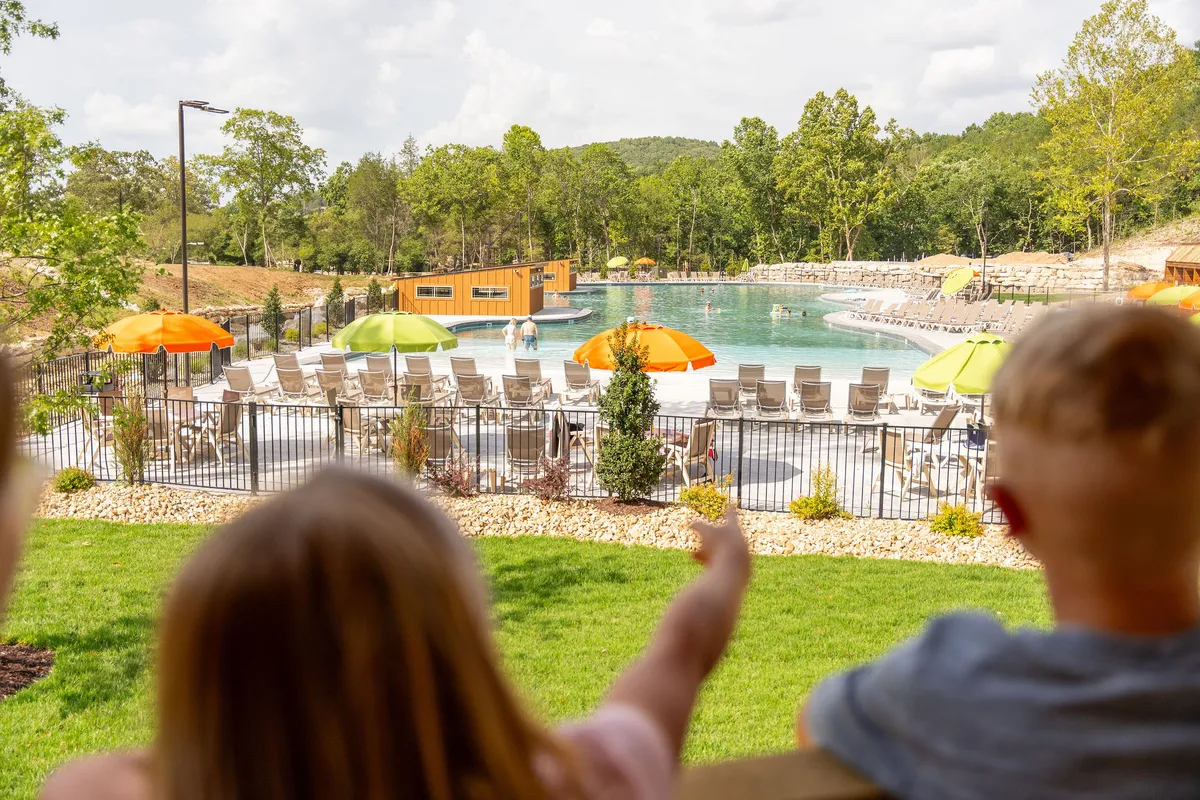 A sun-filled exterior shot of the resort’s pool area, with colorful umbrellas providing shade and children pointing with excitement. The inviting pool is the focal point for swimming, splashing, and sunbathing, all set in a cheerful, energetic environment.