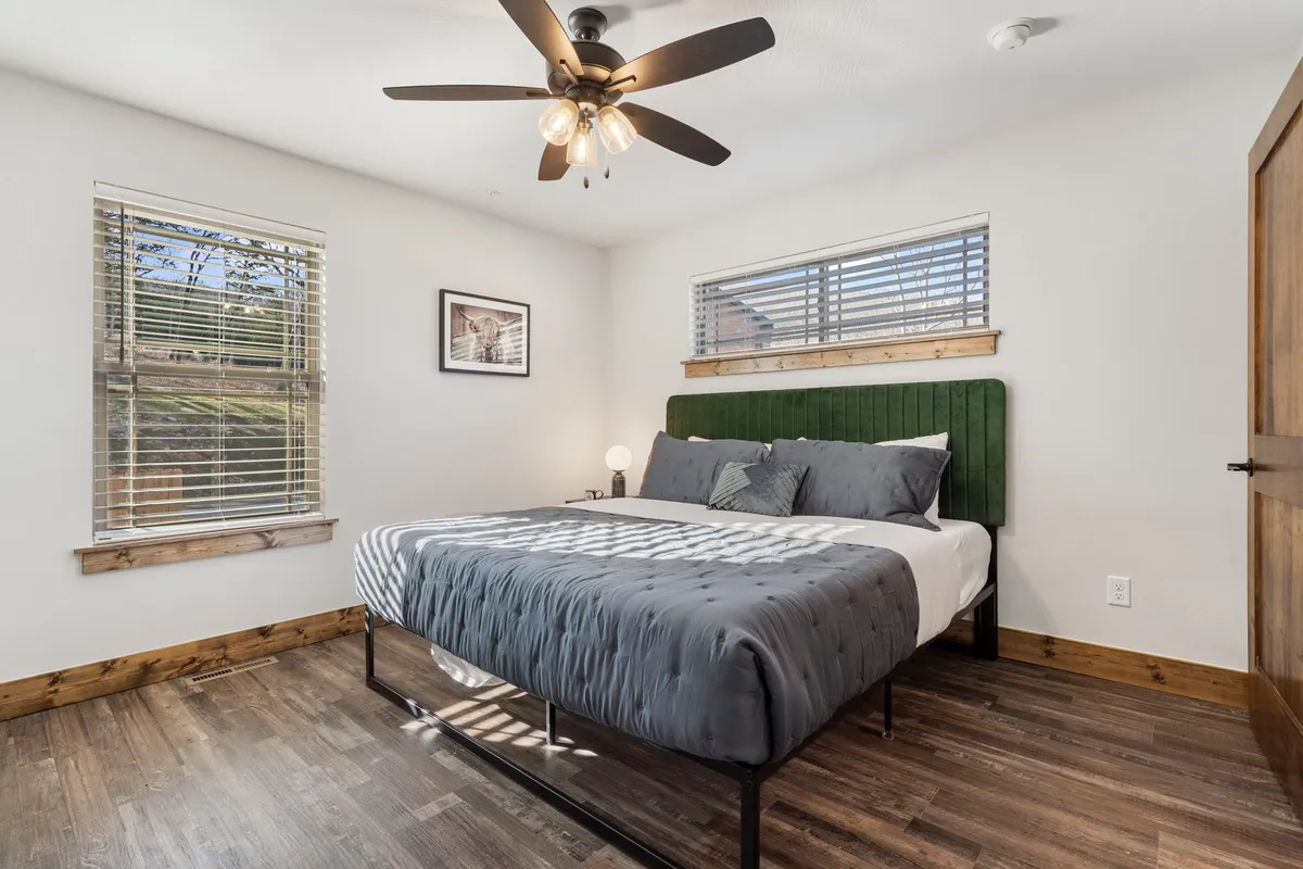 A calm and cozy bedroom with a green upholstered headboard, large window letting in natural light, wooden flooring, and a ceiling fan overhead. The setting is designed for comfort, with space for reading, conversing, or simply unwinding.