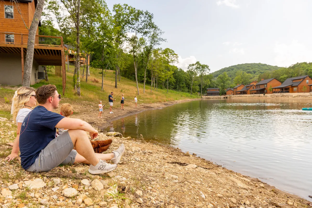 A family sits by the water's edge as children play, with cabins in the background and a natural lakeshore setting. The landscape features calm water and lush greenery, evoking the classic Table Rock Lake experience.