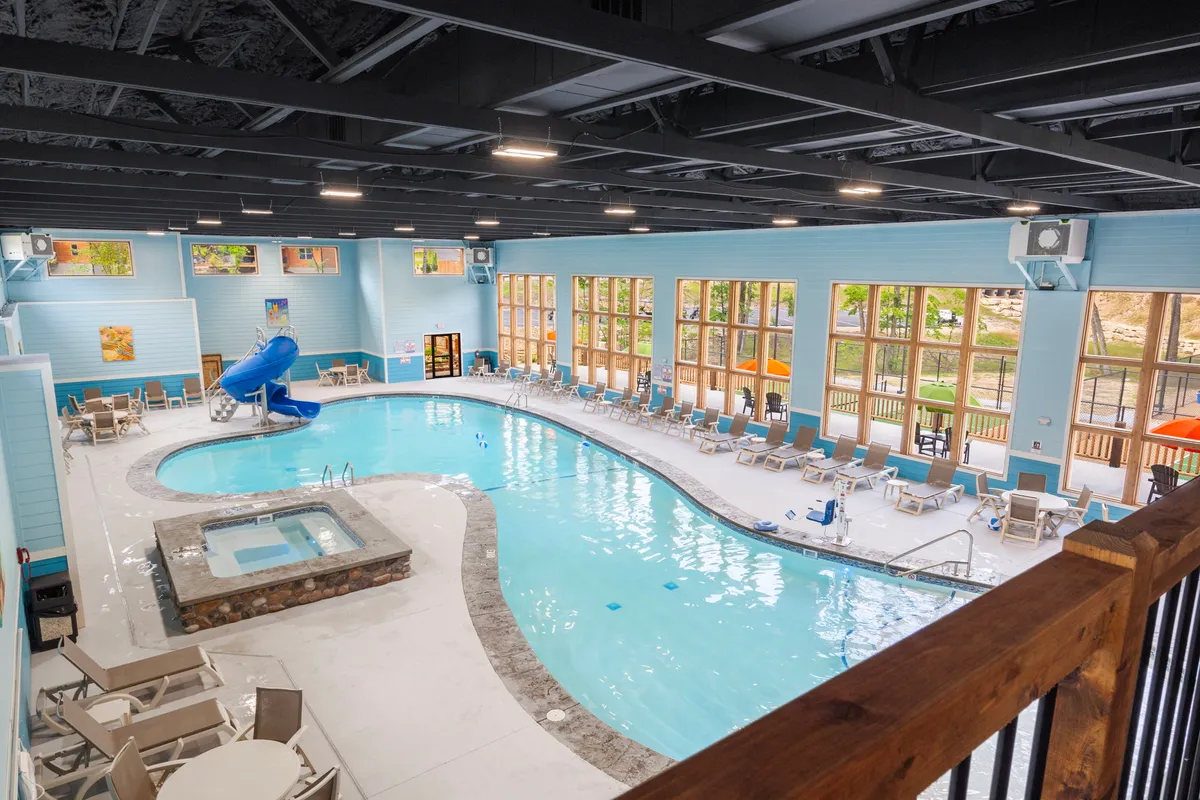 An indoor swimming pool area featuring a water slide, hot tub, and large windows with sunlight streaming in. People can be seen swimming and relaxing, creating a lively atmosphere that feels fun and family-friendly.