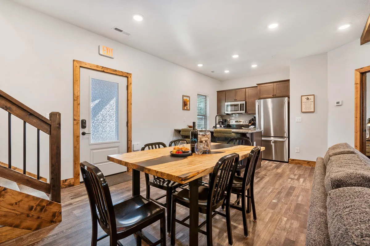 A warm, spacious interior featuring a dining table set adjacent to an open kitchen area. The space is well-lit, with a view of the kitchen and front door, suggesting an inviting atmosphere for shared meals and conversations.
