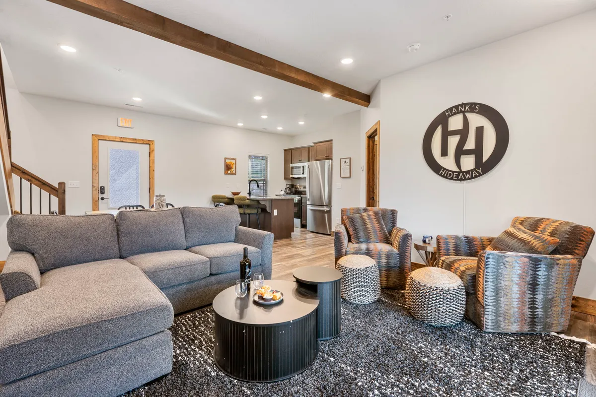 The image showcases a warm and inviting living room inside the cabin, featuring plush sofas, stylish armchairs, a central coffee table, and tasteful wall decor. The cozy setup offers the perfect spot for guests to unwind, socialize, or enjoy a relaxing evening after exploring local attractions.