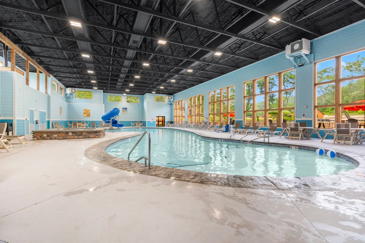 An indoor swimming pool area with a water slide, large windows, and poolside lounge chairs. The mood is joyful and relaxed, with the space clearly designed for family enjoyment rain or shine.