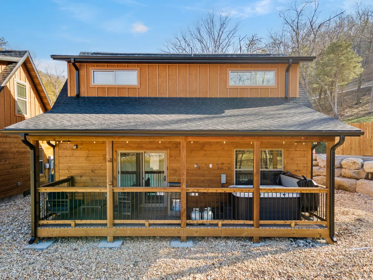 A spacious wooden deck with comfortable outdoor seating looks out over the natural surroundings. Large windows and a stone wall add a touch of rustic luxury, while the setting sun casts a warm glow.