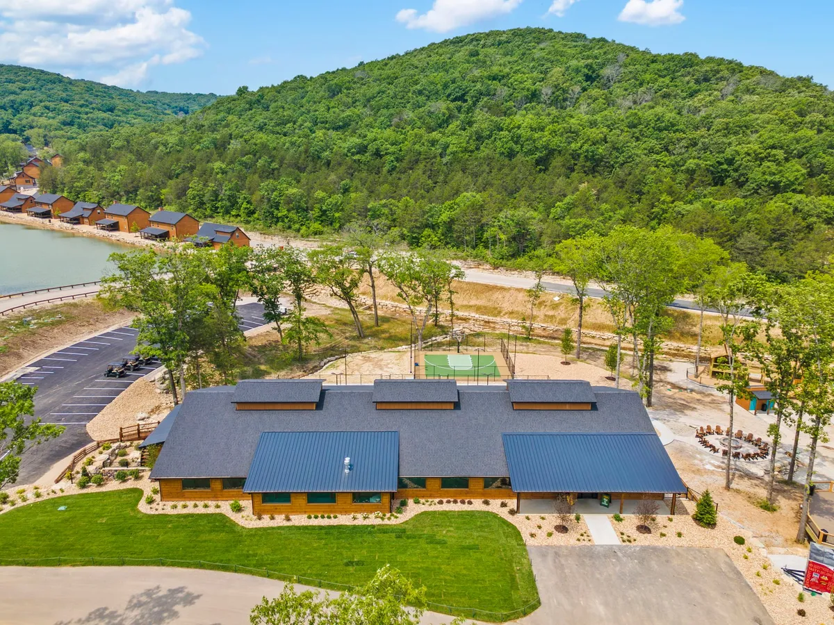 A wide view of the main resort grounds featuring the main building, a waterfront area, basketball court, and a play area. Families engage in a variety of activities, from sports to picnics, surrounded by natural beauty.