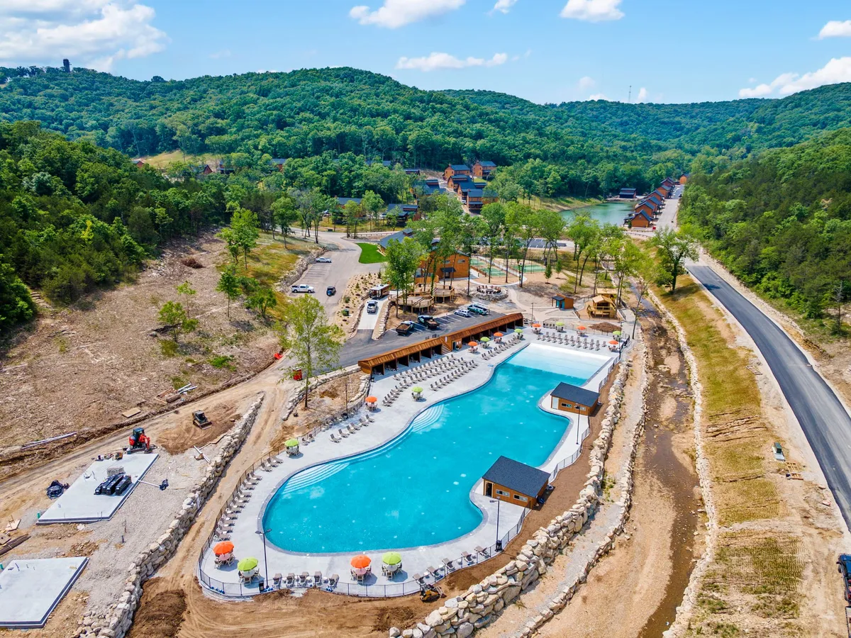 The scene shows a large outdoor resort-style swimming pool with surrounding lounge chairs, cabanas, and nearby cabins set among lush greenery. The setting is vibrant and welcoming, inviting guests to swim or socialize by the poolside.