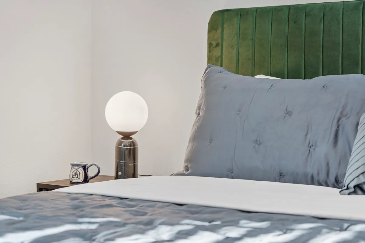 A serene bedroom scene featuring a green velvet headboard, round table lamp, and plush pillows atop the bed. Natural light gently illuminates the room, creating a cozy and inviting environment ideal for relaxation and restful sleep.